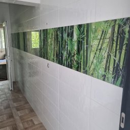 Adrian Bobola - Korytarz wyłożony białymi, prostokątnymi płytkami z poziomym pasem dekoracyjnym z motywem bambusowego lasu; podłoga wyłożona płytkami imitującymi drewno; widoczne drzwi i okno.