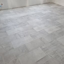 Adrian Bobola - Ułożona, szara podłoga z płytek imitujących kamień w pomieszczeniu w trakcie remontu, widoczne kable wystające ze ściany i częściowo wykończone ściany.