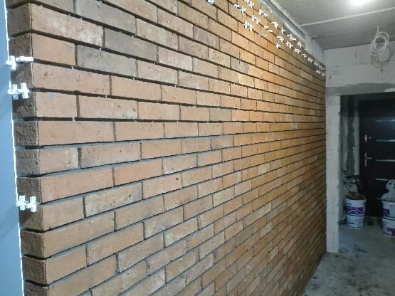Ściana z cegły klinkierowej w trakcie montażu, widoczne krzyżyki dystansowe i puste spoiny, surowy stan wykończenia wnętrza.