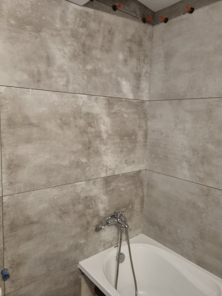 Montaż dużych, szarych płytek imitujących beton w łazience z widoczną wanną i baterią, z użyciem poziomowania krzyżykami i sznurka.