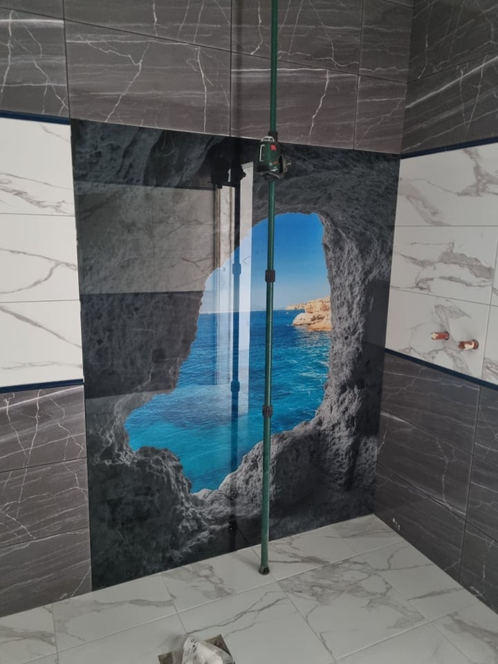 Ściana w łazience wyłożona płytkami imitującymi marmur, z centralnie umieszczoną płytą z fotorealistycznym nadrukiem przedstawiającym morski krajobraz z jaskinią; pionowa rozpórka laserowa...