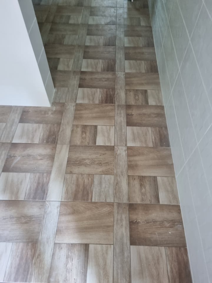 Ułożona podłoga z płytek imitujących drewno w odcieniach brązu i beżu, wzór szachownicy, jasne fugi, fragment białej ściany i szare płytki na ścianie w tle.