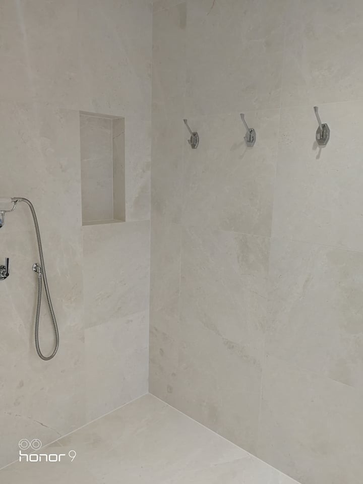 Wykończona kabina prysznicowa z beżowymi płytkami imitującymi marmur, wnęką na kosmetyki i trzema chromowanymi haczykami na ścianie.