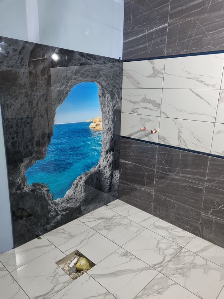 Ściana łazienki wyłożona płytkami z motywem morskim z widokiem na skalną grotę i błękitne morze, obok ściana z szarymi i białymi płytkami imitującymi marmur, podłoga z białych płytek marmurowych...