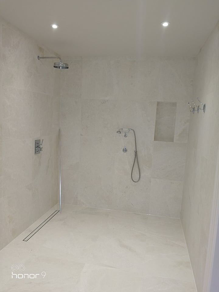 Nowoczesna kabina prysznicowa wyłożona jasnymi, marmurkowymi płytkami, z odpływem liniowym w podłodze, deszczownicą i dodatkową słuchawką prysznicową, wnęką w ścianie i chromowanymi haczykami...