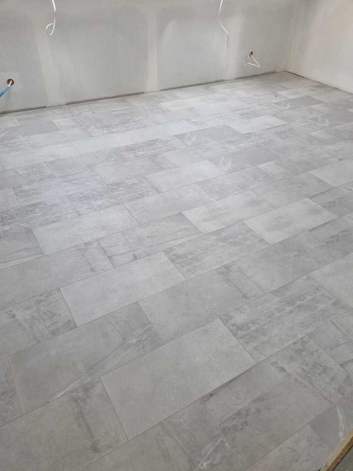 Ułożona, szara podłoga z płytek imitujących kamień w pomieszczeniu w trakcie remontu, widoczne kable wystające ze ściany i częściowo wykończone ściany.