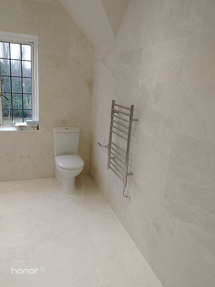 Wykończona łazienka w jasnych, marmurkowych płytkach, z wiszącym grzejnikiem drabinkowym i toaletą kompaktową przy oknie.