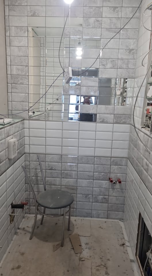 Wnętrze łazienki w trakcie remontu: ściany wyłożone szarymi i białymi płytkami, lustrzana mozaika na górze, widoczne przewody elektryczne, prowizoryczne siedzisko i niedokończona podłoga.