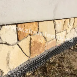 Kamienna elewacja budynku z elementami izolacji pionowej przy gruncie, widoczne nieregularne płyty z piaskowca w odcieniach beżu i brązu, ułożone na fundamencie.