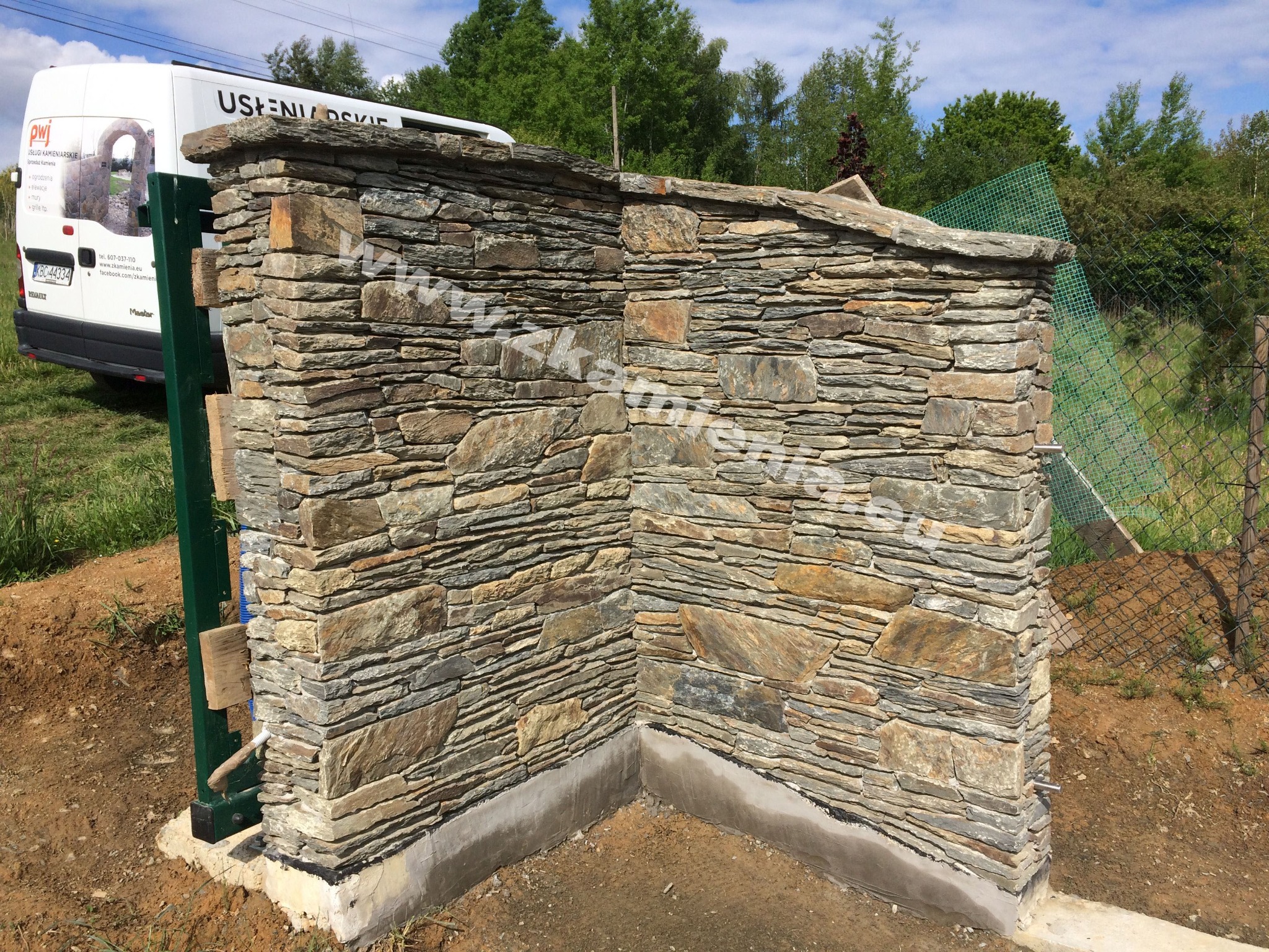 Fragment ogrodzenia z kamienia łupanego w trakcie montażu, widoczna brama z zielonej metalowej ramy i zaparkowany biały bus firmy kamieniarskiej w tle.