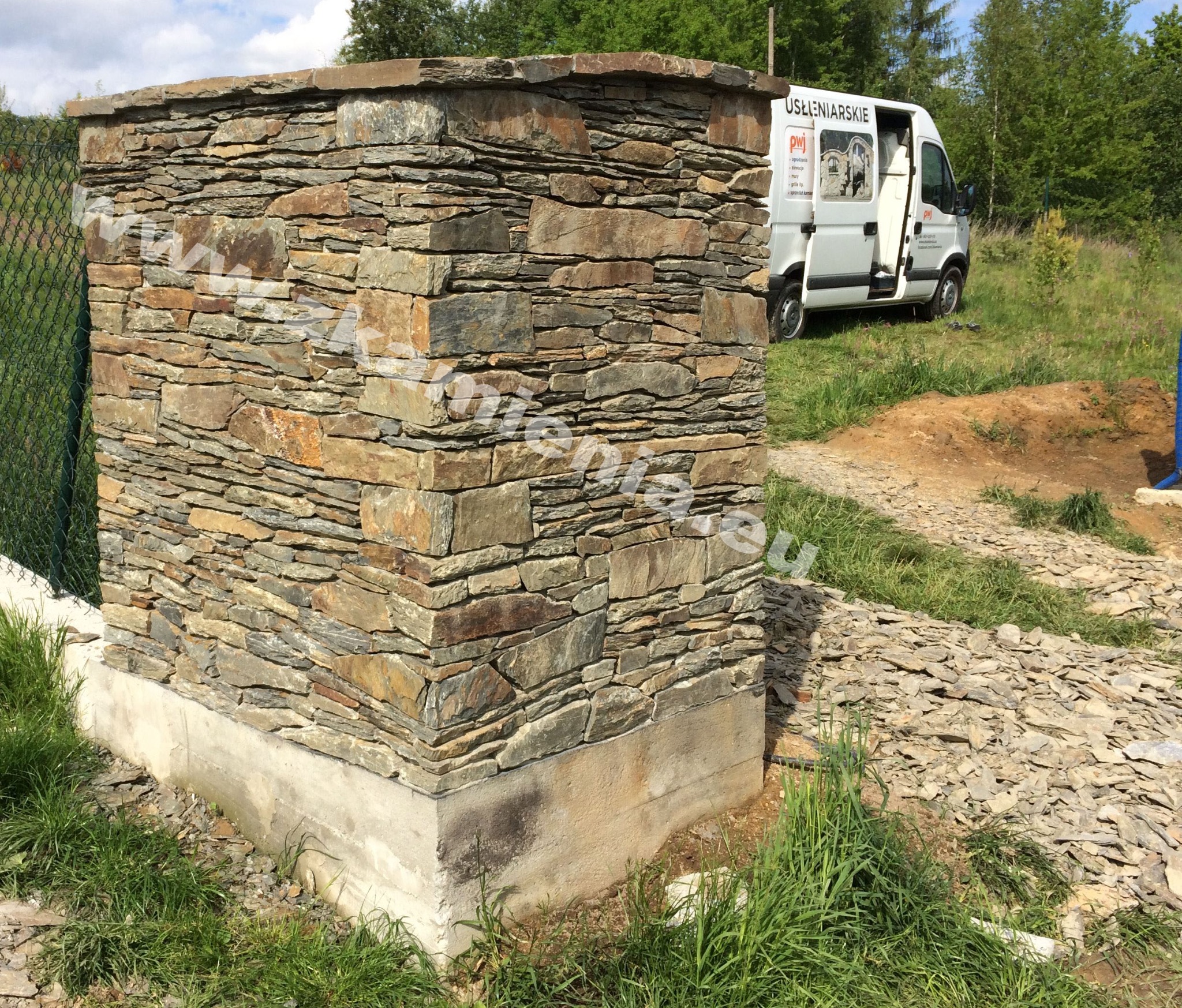 Słupek ogrodzeniowy wykończony nieregularnym kamieniem elewacyjnym, widoczna podmurówka i fragment siatki ogrodzeniowej, w tle zaparkowany biały samochód dostawczy z reklamą firmy.