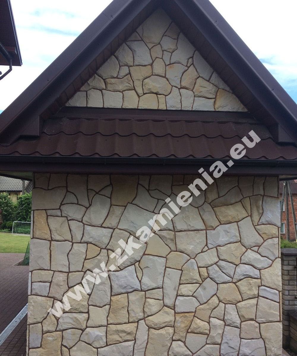 Fragment elewacji budynku wykończony nieregularnymi płytami z jasnego kamienia, widoczny fragment dachu z brązową blachodachówką i orynnowaniem, w tle zieleń trawnika i zabudowania.