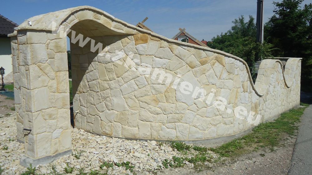 Niski murek z piaskowca z łukowym wejściem i falowaną linią szczytową, otoczony żwirem i trawą, z widocznym domem w tle.