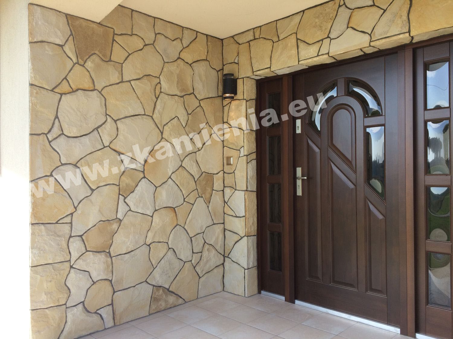 Elewacja budynku wykończona nieregularnym kamieniem w odcieniach beżu i brązu, z ciemnobrązowymi drzwiami wejściowymi z przeszkleniami i nowoczesnym oświetleniem zewnętrznym.