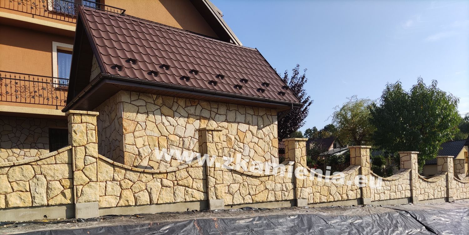 Wykończony murem z kamienia łupanego fragment ogrodzenia z filarami, z widocznym fragmentem domu z brązowym dachem i balkonem z ozdobną balustradą, zabezpieczony folią fundament.
