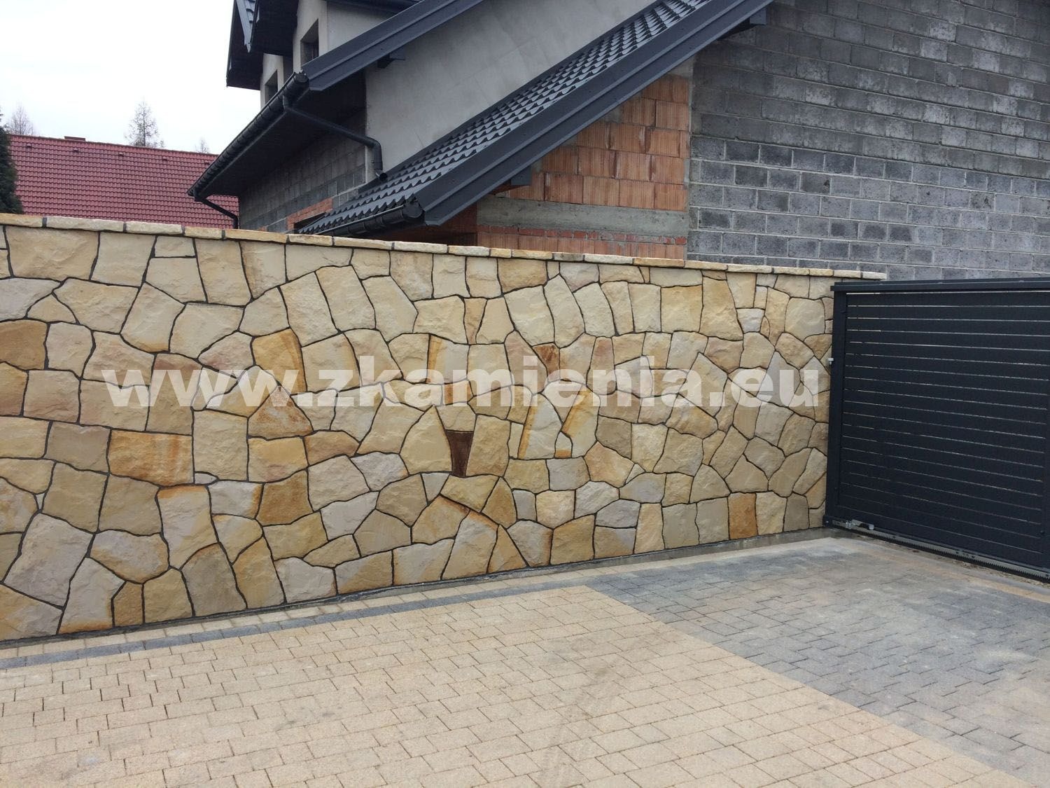 Kamienny mur ogrodzeniowy z nieregularnych, piaskowych płyt, przylegający do czarnej, metalowej bramy przesuwnej i budynku z cegły oraz szarego bloczka. Na kamiennej powierzchni widoczny znak wodny...