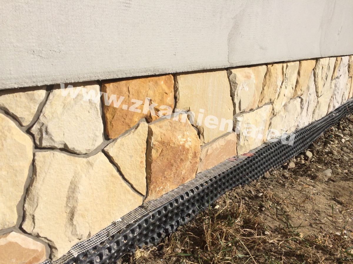 Kamienna elewacja budynku z elementami izolacji pionowej przy gruncie, widoczne nieregularne płyty z piaskowca w odcieniach beżu i brązu, ułożone na fundamencie.