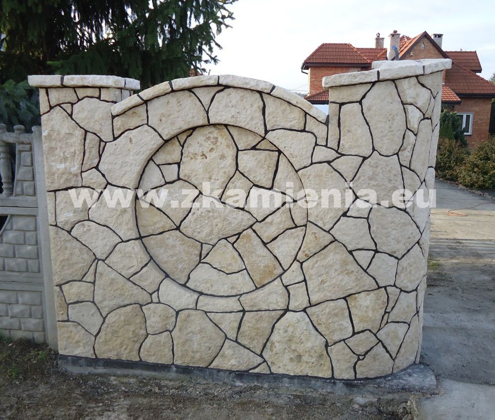Fragment ogrodzenia z nieregularnych, jasnych kamieni z ciemnymi fugami, z okrągłym wzorem, przylegający do betonowego ogrodzenia, na tle domu z czerwoną dachówką.