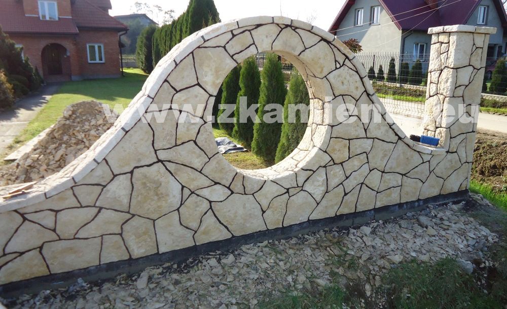 Nietypowe ogrodzenie z piaskowca o nieregularnych kształtach, z okrągłym otworem, w tle widoczne tuje i domy.