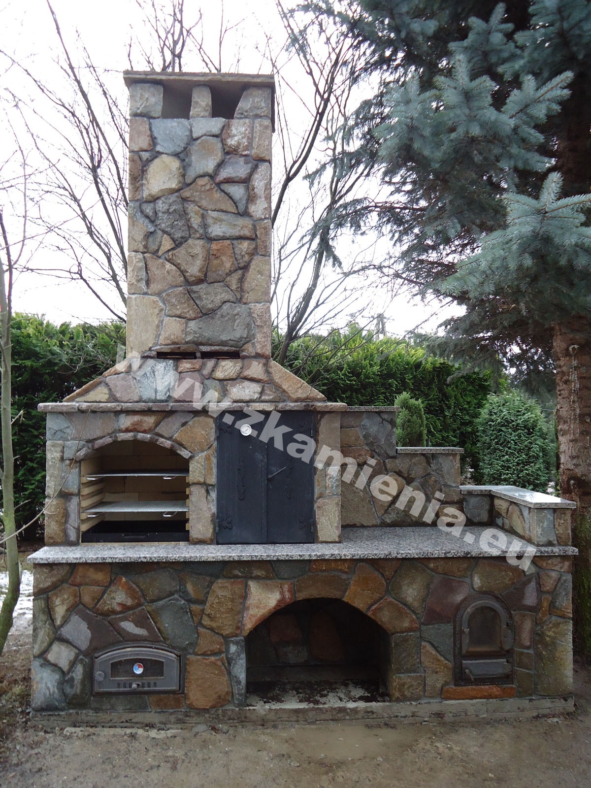 Kamienny grill ogrodowy z piecem i wędzarnią, wykonany z nieregularnych kamieni w odcieniach beżu i szarości, z czarnymi drzwiczkami i kominem, na tle ogrodu z drzewami iglastymi.