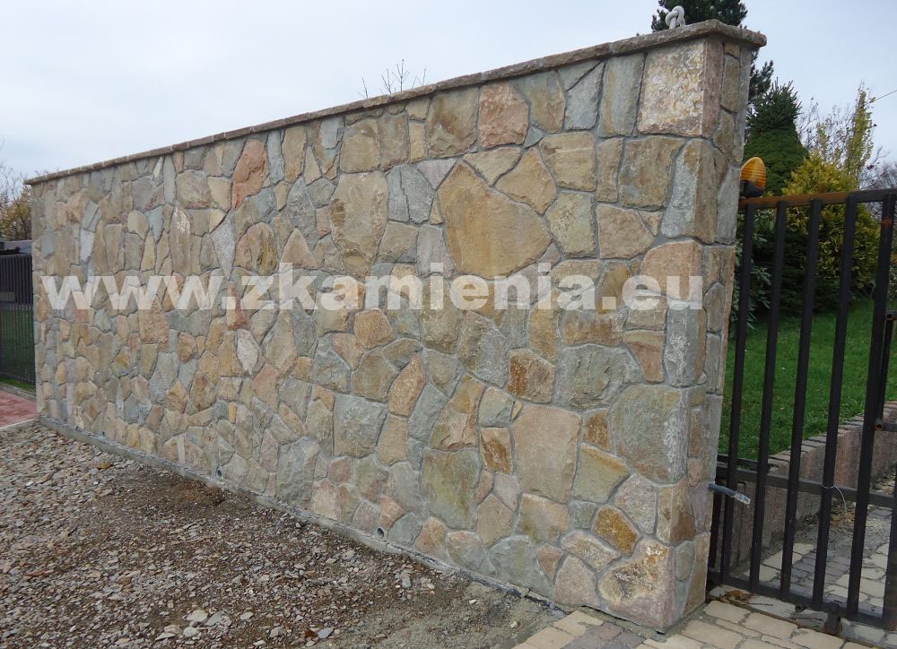 Niski mur z nieregularnych kamieni w odcieniach beżu i szarości, z widoczną bramą wejściową i fragmentem ogrodu w tle, z naniesionym adresem strony internetowej.