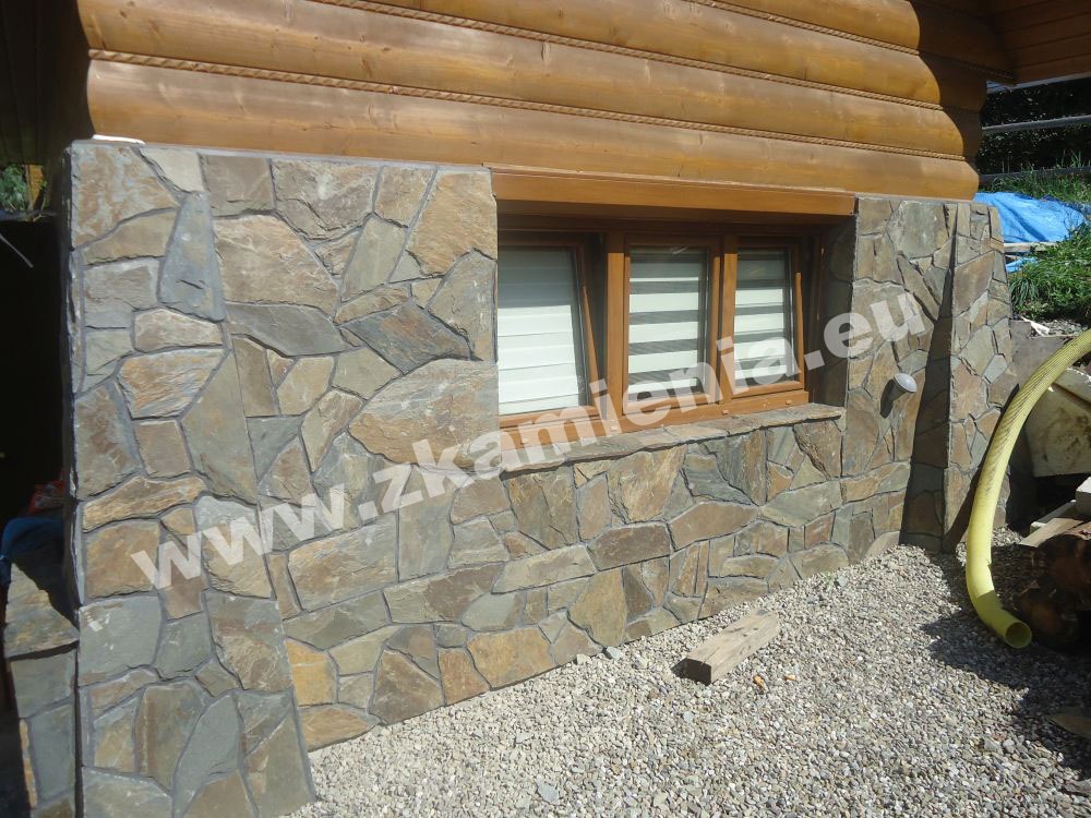 Kamienna elewacja budynku z oknem, wykonana z nieregularnych, szarobrązowych płytek, z widocznym adresem strony internetowej na fasadzie.