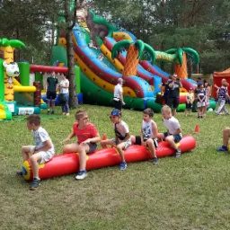 Dzieci siedzące na dmuchanych wałkach podczas zabawy na świeżym powietrzu, w tle dmuchane zamki i zjeżdżalnie w motywie dżungli.