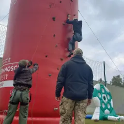 Mężczyzna wspina się na dmuchaną ściankę wspinaczkową, zabezpieczany liną przez kobietę w uprzęży; w tle mężczyzna w spodniach moro obserwuje, a obok widoczna dmuchana tarcza do kopania piłek.