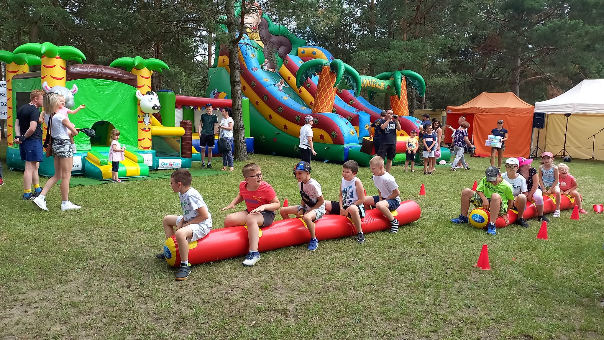 Dzieci siedzące na dmuchanych wałkach podczas zabawy na świeżym powietrzu, w tle dmuchane zamki i zjeżdżalnie w motywie dżungli.