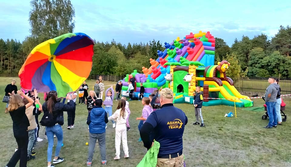 Dzieci bawią się na zewnątrz z kolorową chustą animacyjną i dmuchanym zamkiem w tle.