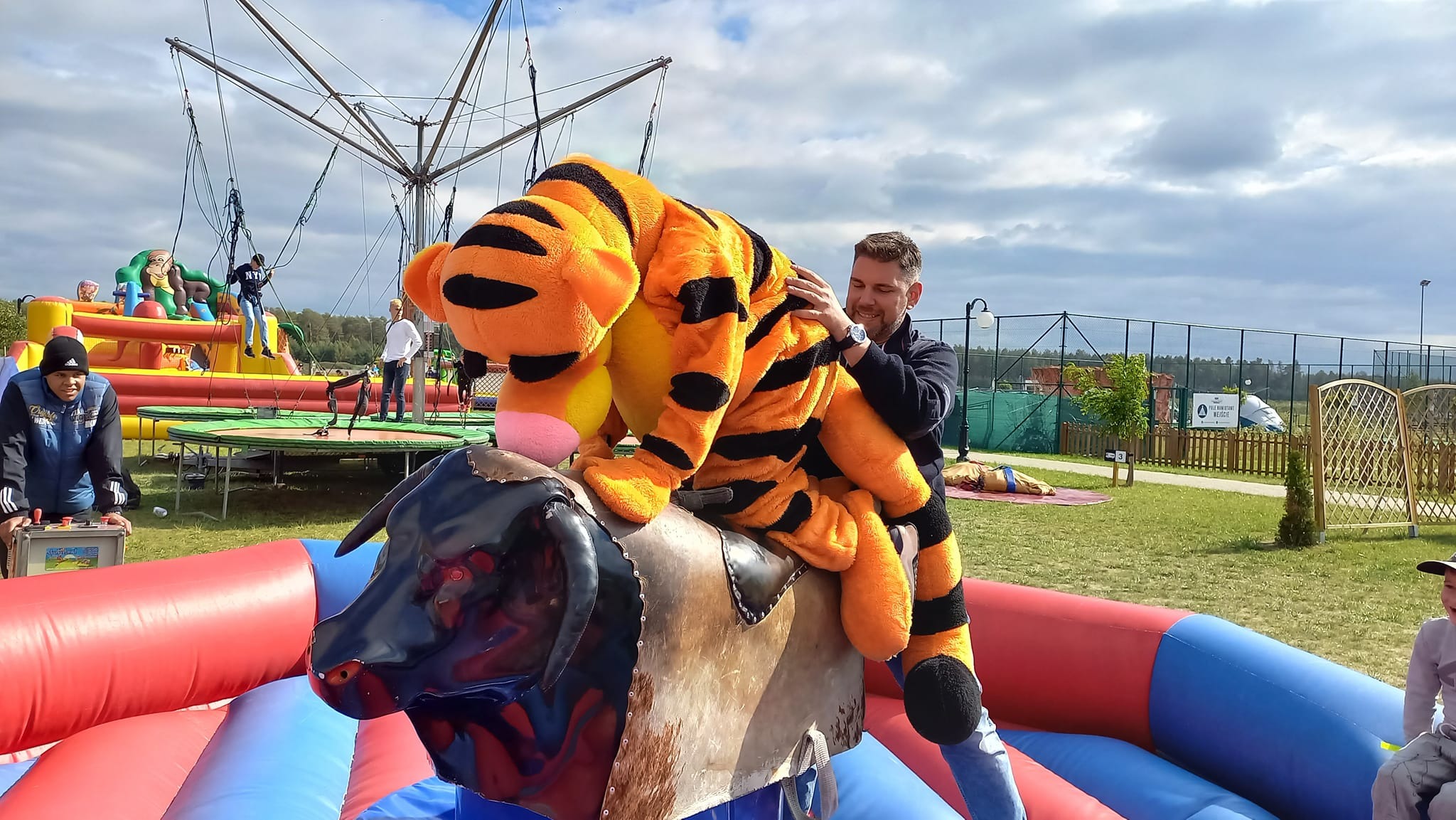 Uśmiechnięty mężczyzna pomaga dziecku w kostiumie tygrysa utrzymać się na mechanicznym byku na tle dmuchanego zamku i trampoliny.