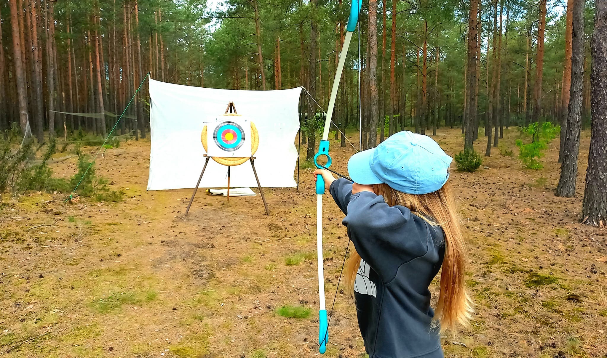 Dziewczynka w niebieskiej czapce naciąga cięciwę łuku, celując w tarczę na tle leśnej scenerii.