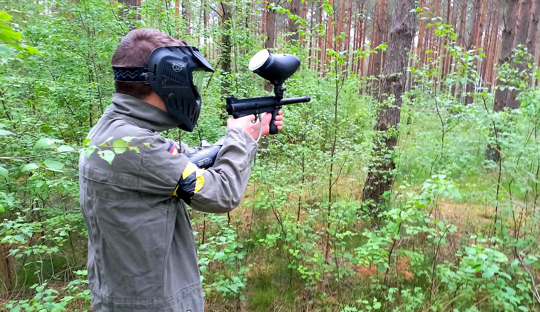 Osoba w masce ochronnej i wojskowej kurtce celuje z pistoletu paintballowego w lesie.