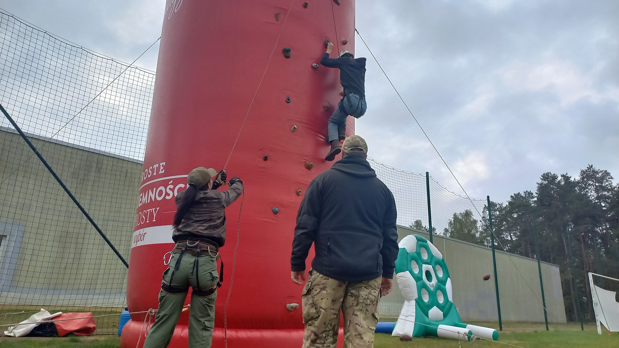 Mężczyzna wspina się na dmuchaną ściankę wspinaczkową, zabezpieczany liną przez kobietę w uprzęży; w tle mężczyzna w spodniach moro obserwuje, a obok widoczna dmuchana tarcza do kopania piłek.