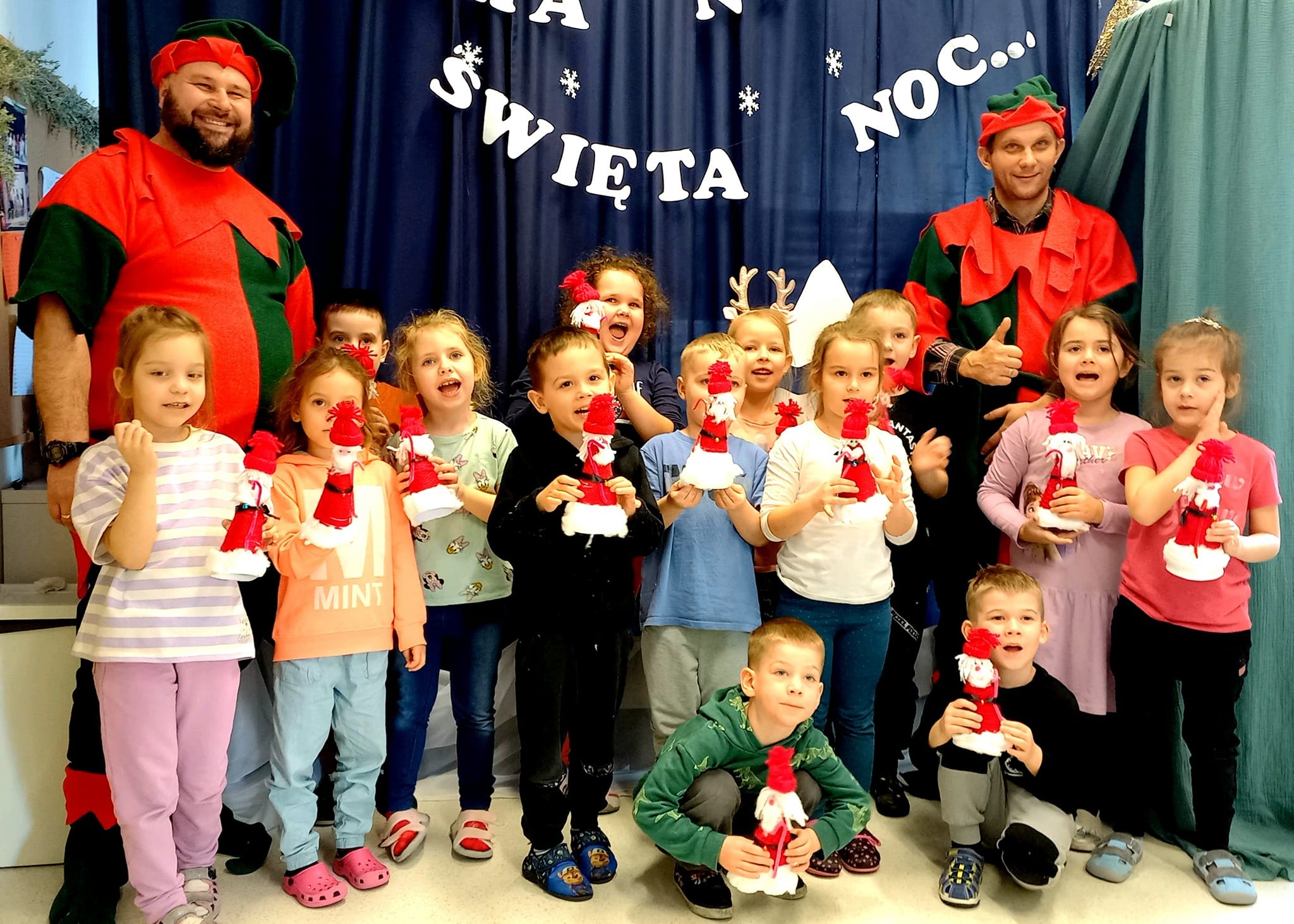 Grupa dzieci z animatorami przebranymi za elfy trzyma własnoręcznie zrobione, świąteczne ozdoby na tle dekoracji z napisem 'Święta Noc...'