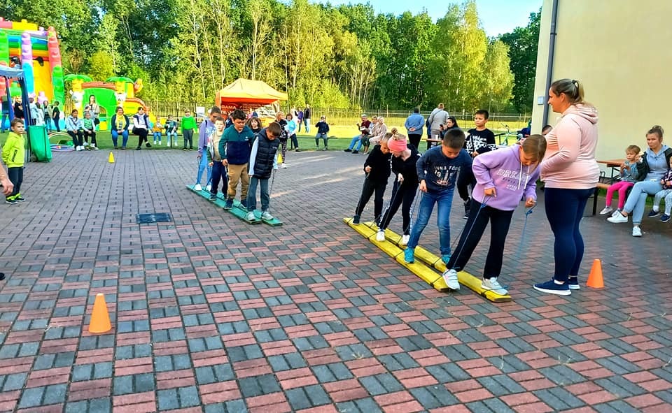 Grupa dzieci uczestniczy w zabawie zespołowej na drewnianych nartach podczas imprezy plenerowej, w tle dmuchany zamek i siedzący widzowie.