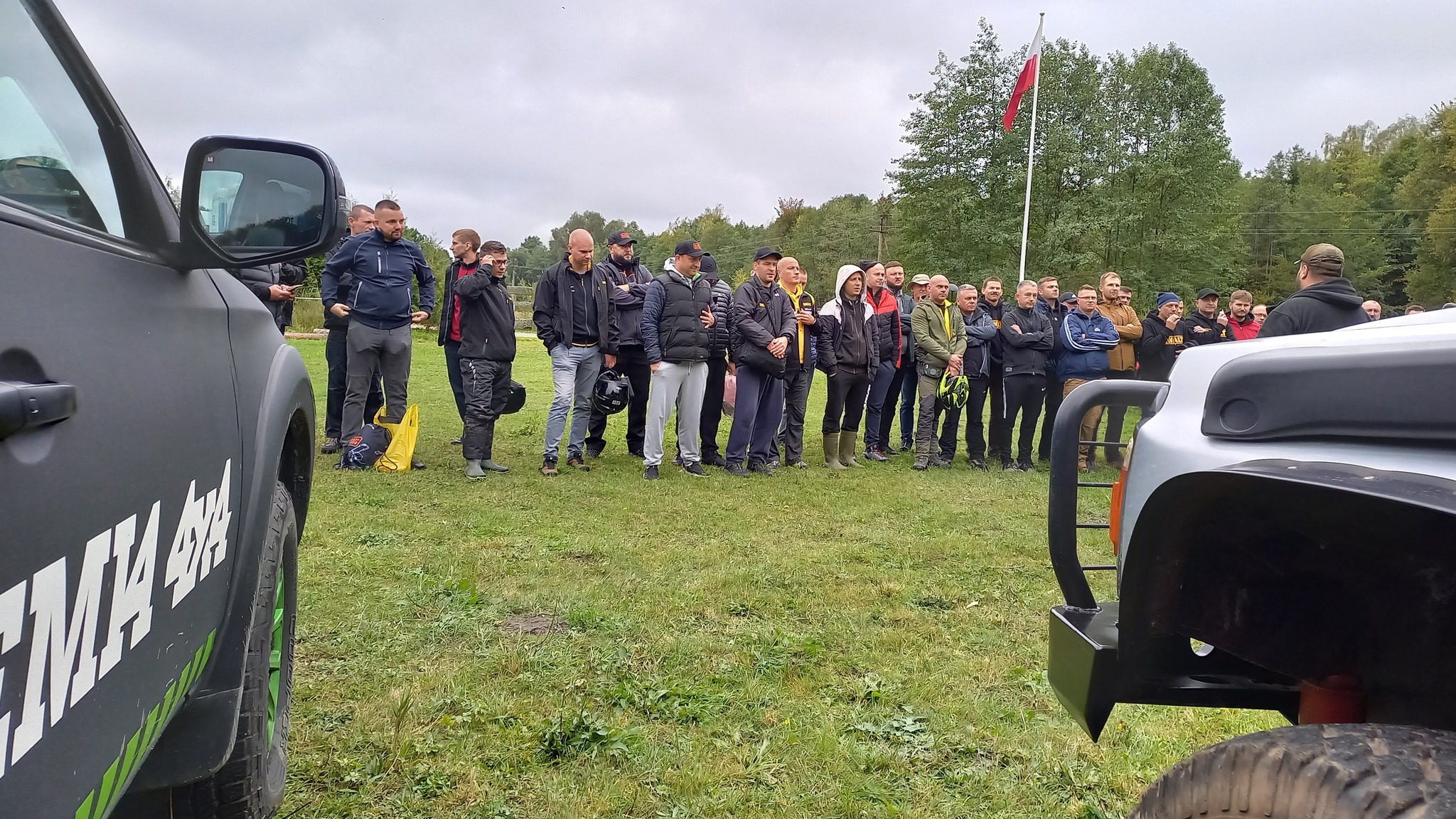 Grupa osób stojąca na trawie przed dwoma samochodami terenowymi, w tle las i flaga Polski.