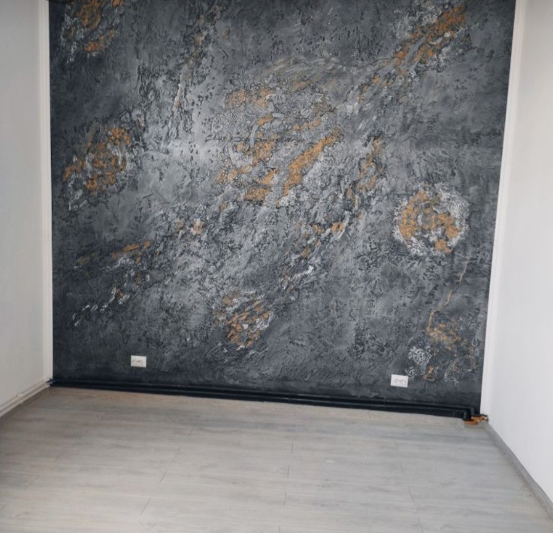 Ściana w pokoju wykończona dekoracyjnym tynkiem w odcieniach szarości z akcentami złota, imitującymi naturalny kamień, widoczne gniazdka elektryczne i drewniana podłoga.