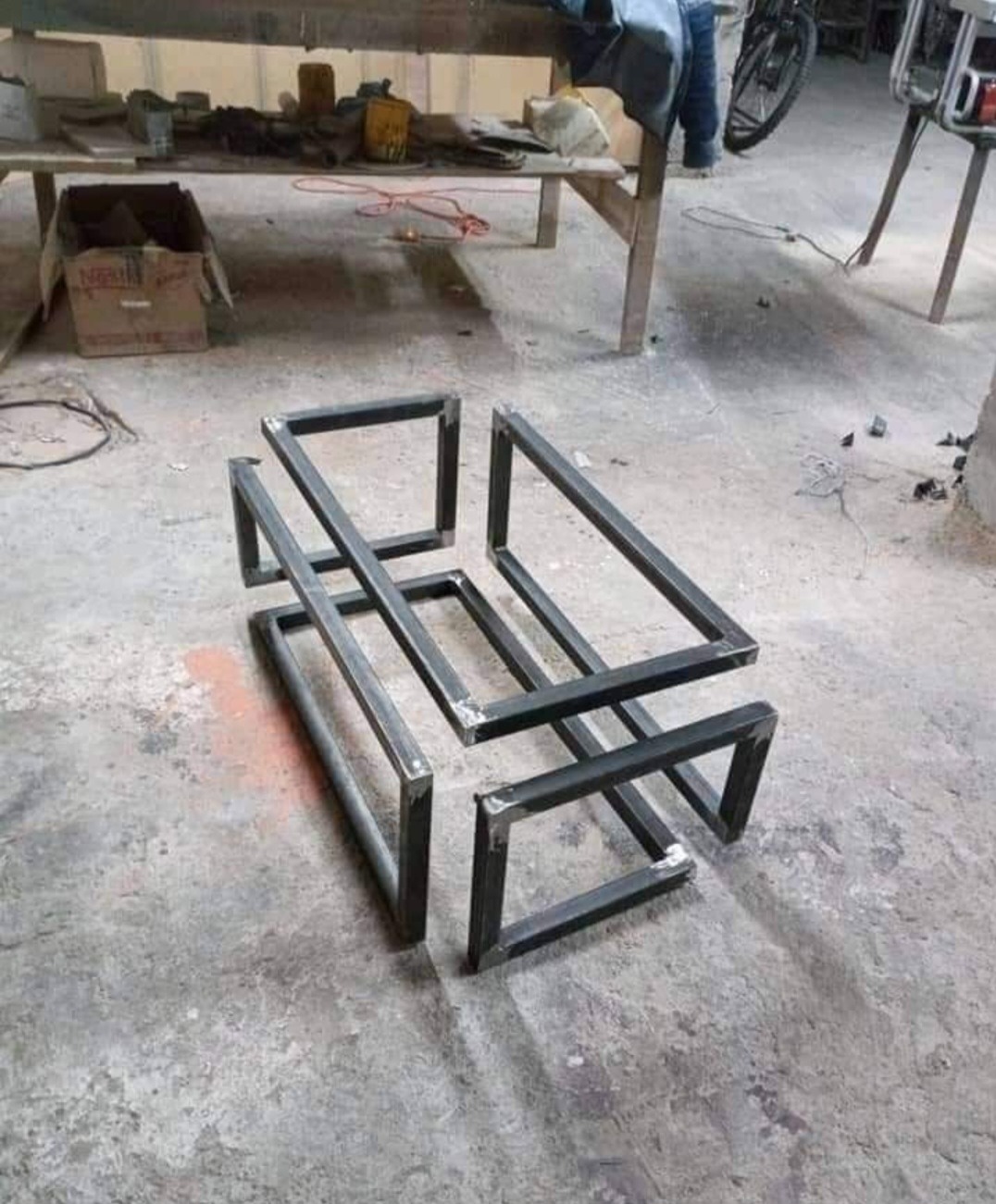 Surowa, spawana konstrukcja z metalowych profili, przypominająca szkielet mebla, leżąca na betonowej podłodze w warsztacie.