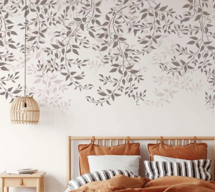 Wzorzysta ściana w sypialni z motywem liści, rattanowa lampa wisząca i drewniane łóżko z pomarańczową pościelą.