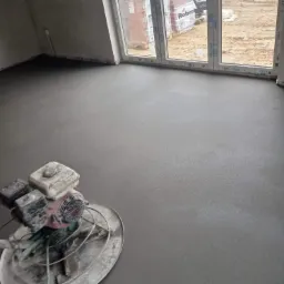Świeżo wylana, gładka posadzka w pomieszczeniu, zacierana mechanicznie zacieraczką do betonu. W tle okno z widokiem na plac budowy i palety z cegłami.