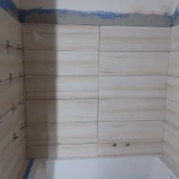 Ściana łazienki w trakcie układania beżowych płytek imitujących drewno, widoczne krzyżyki dystansowe i fragment wanny.