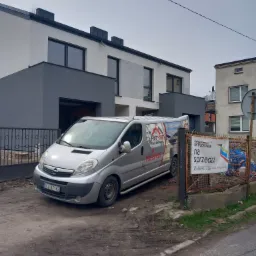 Srebrny bus firmy budowlanej Piotr-Bud zaparkowany przed nowo wybudowanym domem z szaro-białą elewacją, na ogrodzeniu wisi baner z napisem 'na sprzedaż'.