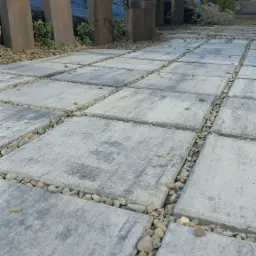 Nowoczesna ścieżka z szarych płyt betonowych, wypełniona drobnymi kamieniami. W tle minimalistyczna zieleń i elementy drewniane, tworzące spójną kompozycję ogrodu.