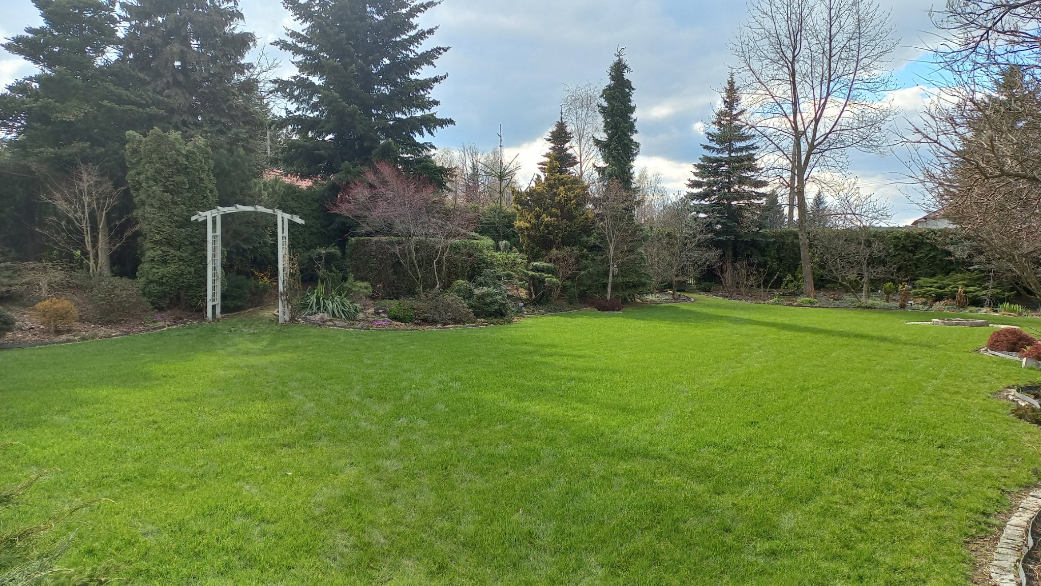 Rozległy, zadbany trawnik z drzewami iglastymi i liściastymi w tle, ozdobiony białą pergolą i rabatami kwiatowymi, tworzący harmonijny krajobraz ogrodu.