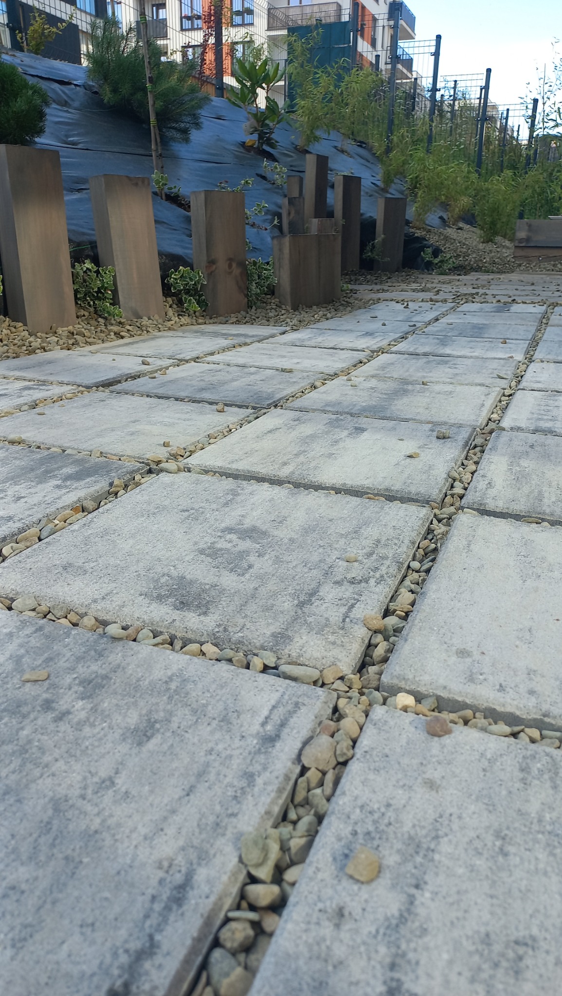 Nowoczesna ścieżka z szarych płyt betonowych, wypełniona drobnymi kamieniami. W tle minimalistyczna zieleń i elementy drewniane, tworzące spójną kompozycję ogrodu.