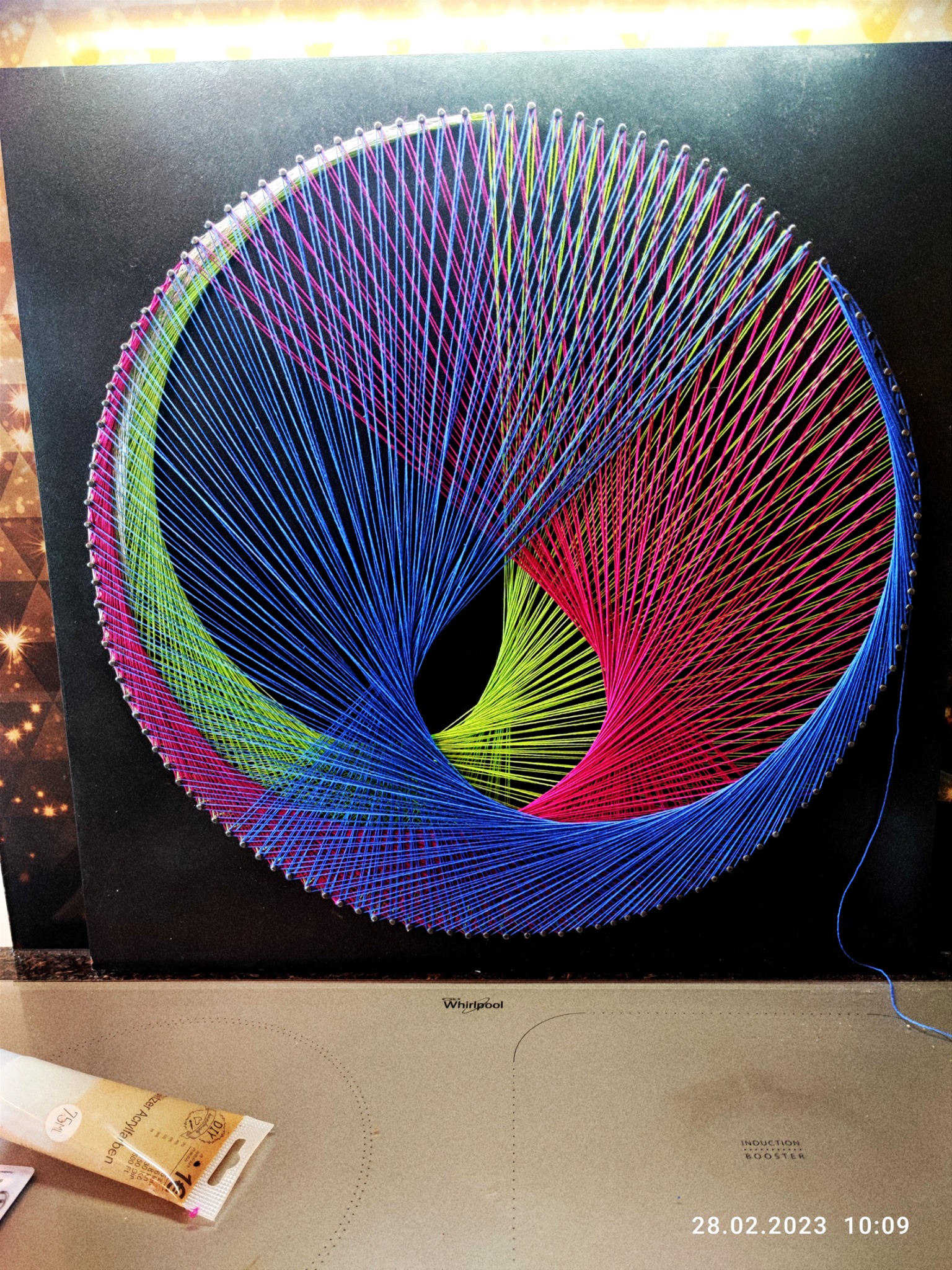 Niedokończona grafika string art na czarnym tle, przedstawiająca abstrakcyjny okrąg z naprzemiennych nici: niebieskich, różowych i zielonych, przymocowanych do szpilek; widoczna tuba farby...