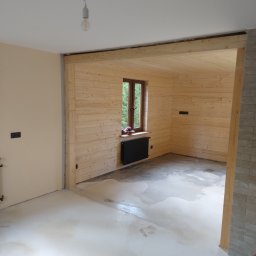 LUK-BUD Łukasz Wołoch - Wnętrze w trakcie remontu: ściany wyłożone jasnym drewnem, wylewka na podłodze, widoczne okno z kwiatami i prowizoryczne oświetlenie żarówką na kablu.