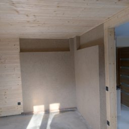 LUK-BUD Łukasz Wołoch - Wnętrze w trakcie prac wykończeniowych: drewniana boazeria na suficie i ścianie, ściana pokryta beżową strukturą, surowa drewniana rama drzwiowa, wylewka na podłodze, promienie słońca wpadające...