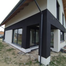 LUK-BUD Łukasz Wołoch - Nowoczesny dom jednorodzinny z elewacją w trakcie wykończenia: czarne pasy i białe fragmenty, widoczne okna i elementy konstrukcyjne, teren wokół domu częściowo uporządkowany.
